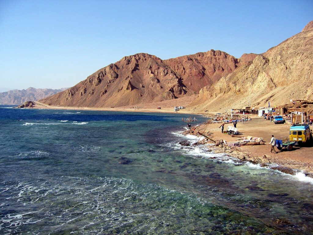 La playa del Blue Hole: los cafés beduinos al pie de las montañas ocres, algunos vehículos y el mar agitado en primer plano