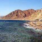 La playa del Blue Hole: los cafés beduinos al pie de las montañas ocres, algunos vehículos y el mar agitado en primer plano