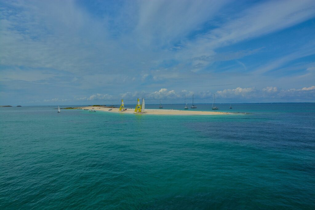 Una larga lengua de arena blanca donde están varados catamaranes de velas amarillas y blancas, rodeada de veleros fondeados en un agua turquesa, bajo un cielo azul surcado de nubes.