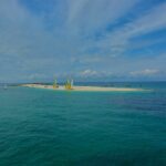 Una larga lengua de arena blanca donde están varados catamaranes de velas amarillas y blancas, rodeada de veleros fondeados en un agua turquesa, bajo un cielo azul surcado de nubes.