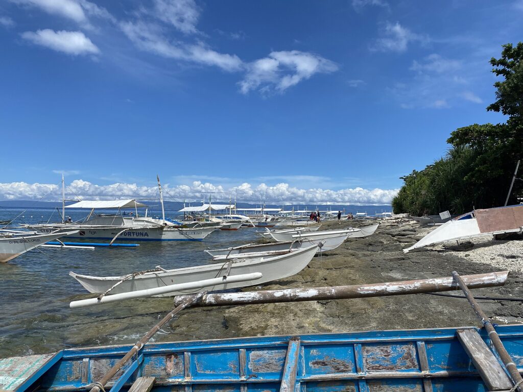 Una fila de bangkas blancas amarradas a lo largo de una orilla rocosa, pequeñas canoas con balancín varadas sobre la roca y una barca azul desconchada en primer plano, bajo un cielo azul con nubes blancas.