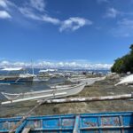 Una fila de bangkas blancas amarradas a lo largo de una orilla rocosa, pequeñas canoas con balancín varadas sobre la roca y una barca azul desconchada en primer plano, bajo un cielo azul con nubes blancas.