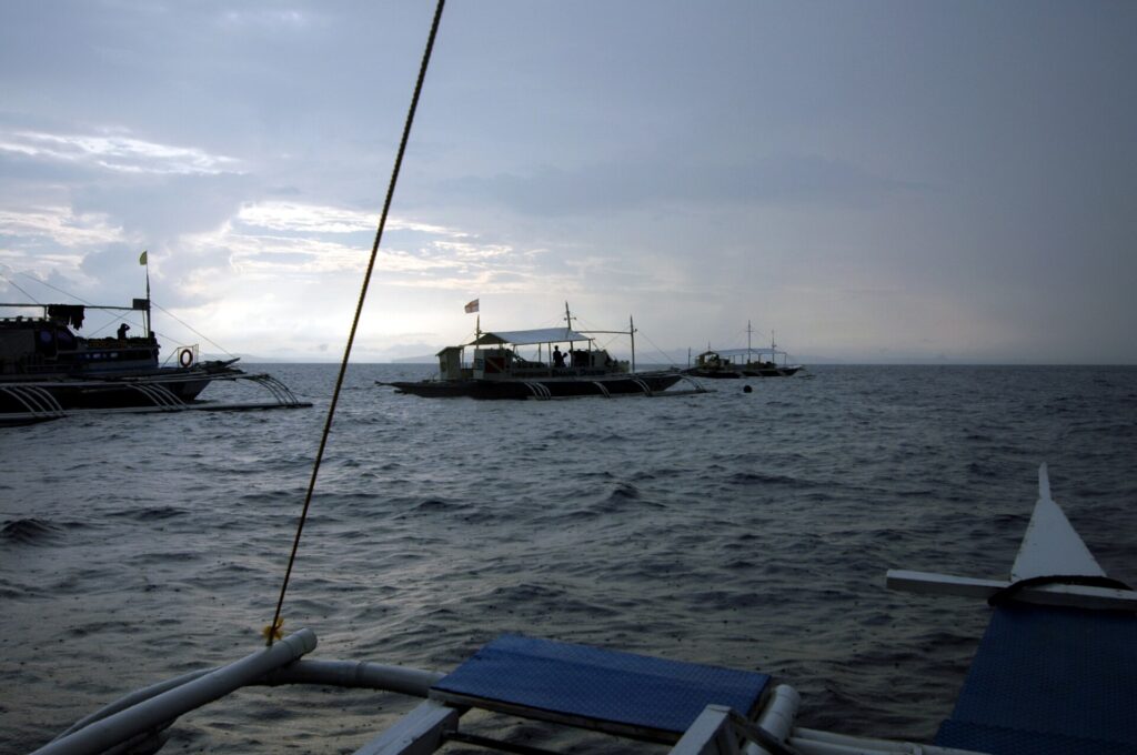 Tres bancas de buceo fondeadas en Monad Shoal bajo un cielo gris de madrugada, vistas desde la cubierta de otro barco