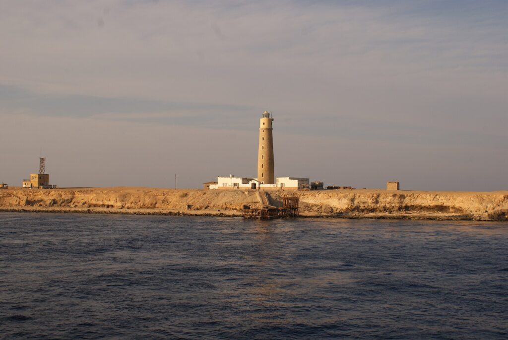 El faro de Big Brother y sus edificios blancos en el islote desértico, con un embarcadero oxidado en primer plano, a la luz dorada de la tarde