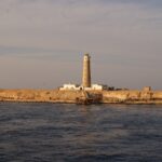 El faro de Big Brother y sus edificios blancos en el islote desértico, con un embarcadero oxidado en primer plano, a la luz dorada de la tarde