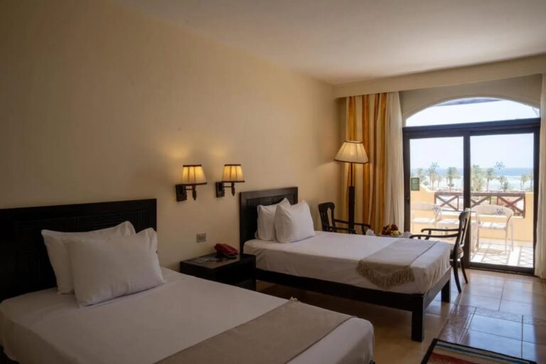 Una habitación con dos camas abierta a un balcón, con vista a las palmeras y al mar
