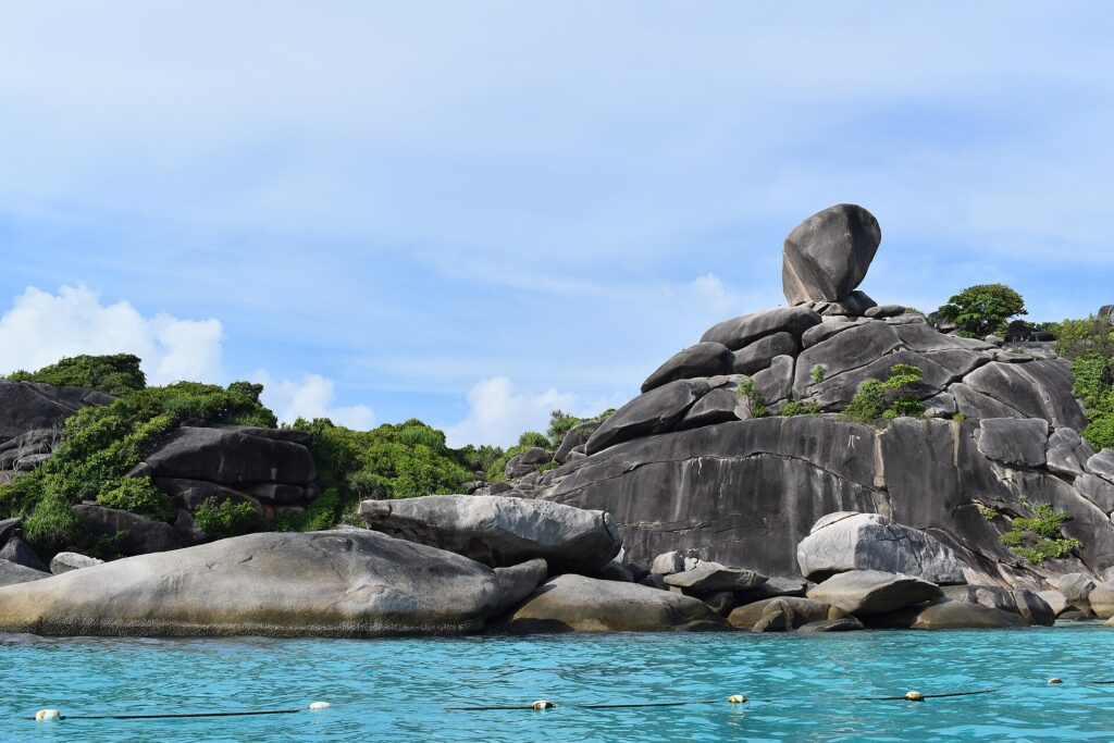 El enorme bloque de granito en equilibrio de la isla 8 corona un montón de rocas grises y lisas al borde de un agua turquesa, bajo un cielo azul velado, con una línea de boyas en primer plano