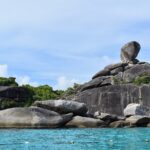 El enorme bloque de granito en equilibrio de la isla 8 corona un montón de rocas grises y lisas al borde de un agua turquesa, bajo un cielo azul velado, con una línea de boyas en primer plano