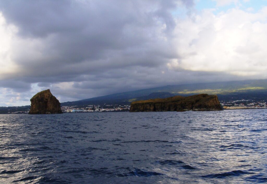 Vistos desde un barco sobre un mar oscuro y agitado, un islote rocoso erguido a la izquierda y un islote largo y plano a la derecha, frente a la ciudad de Madalena y las laderas de Pico bajo un cielo cargado