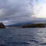 Vistos desde un barco sobre un mar oscuro y agitado, un islote rocoso erguido a la izquierda y un islote largo y plano a la derecha, frente a la ciudad de Madalena y las laderas de Pico bajo un cielo cargado