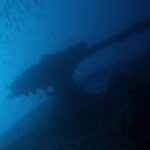 El cañón de popa del Thistlegorm a contraluz sobre el azul, bajo un banco de peces pequeños