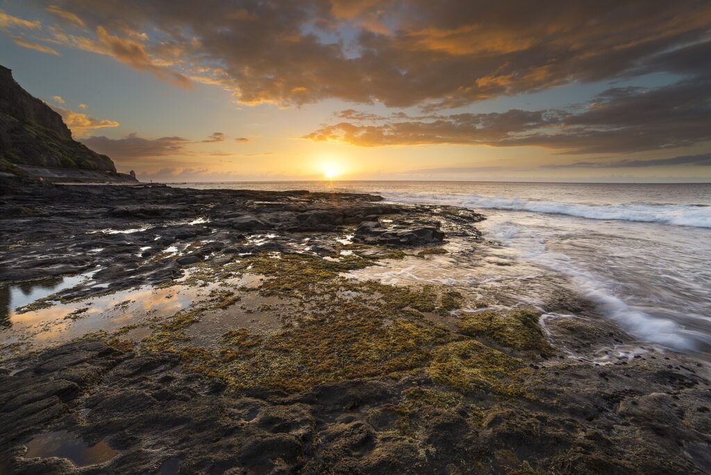 Puesta de sol sobre el océano vista desde una plataforma de basalto negro cubierta de algas y charcas, con las olas que la lamen a la derecha y un acantilado a la izquierda