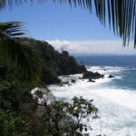 Vista en picado enmarcada por hojas de palmera: un cabo cubierto de selva tropical cae al mar, las olas rompen blancas sobre rocas negras y una costa lejana se dibuja en el horizonte bajo un cielo azul.