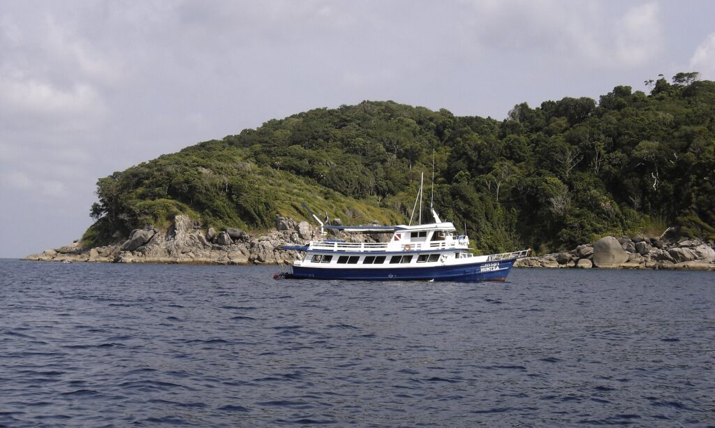 Un barco de crucero azul y blanco fondeado frente a una isla de las Similan, con sus bloques de granito gris a ras del agua bajo la selva