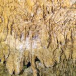 Primer plano de formaciones amarillo pálido y estalactitas finas, con el reflejo ondulado del agua abajo