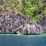 Agujas de caliza gris esculpidas en láminas se hunden en un agua turquesa y luego azul profundo, al pie de una ladera cubierta de bosque.