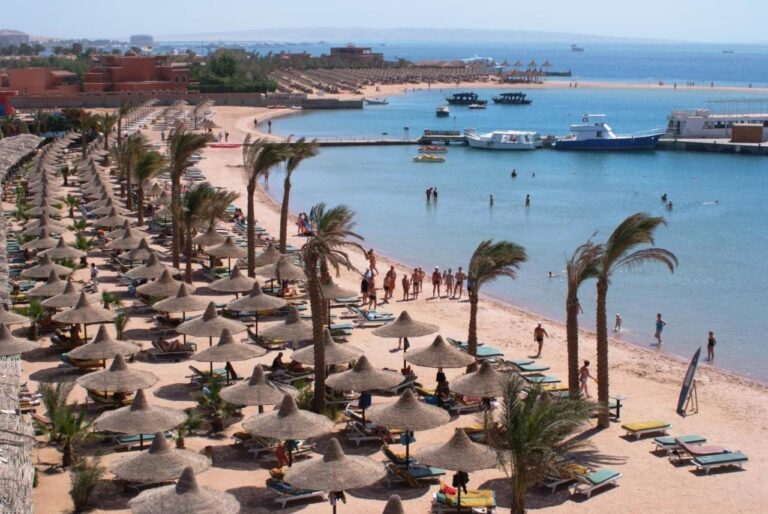 La playa del Giftun Azur Resort, sus sombrillas de paja y sus palmeras, con un muelle y barcos al fondo