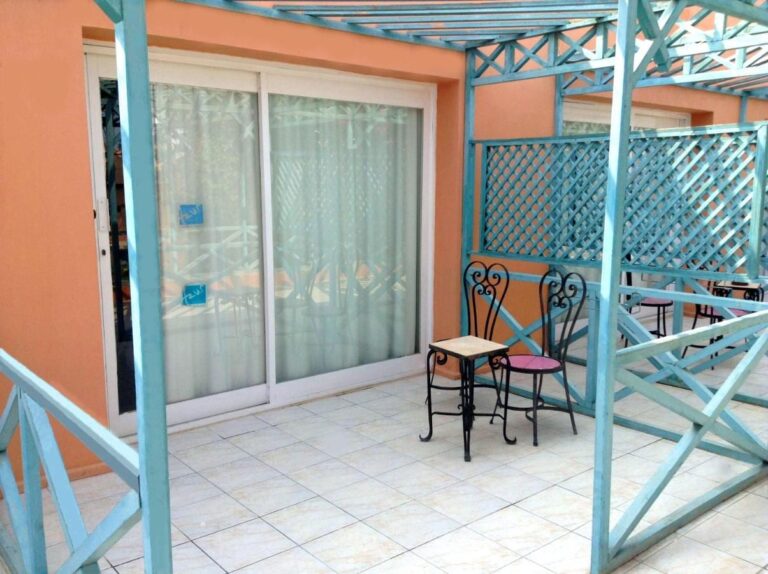 Una terraza embaldosada bajo una pérgola turquesa, con una mesita y dos sillas de hierro forjado
