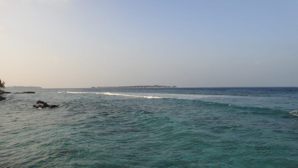 Desde una orilla rocosa, un paso turquesa agitado donde una línea de olas rompe sobre el arrecife, y en el horizonte una isla baja bordeada de bungalós sobre pilotes, bajo un cielo pálido.