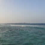 Desde una orilla rocosa, un paso turquesa agitado donde una línea de olas rompe sobre el arrecife, y en el horizonte una isla baja bordeada de bungalós sobre pilotes, bajo un cielo pálido.
