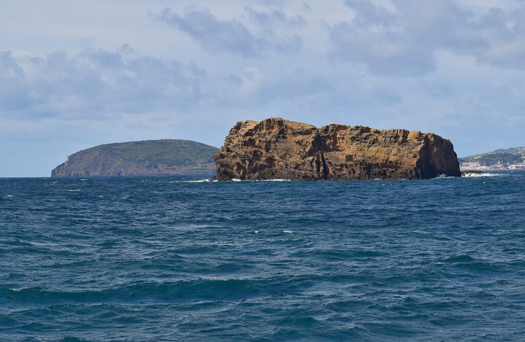 El Ilhéu Deitado, largo bloque de toba ocre con acantilados llenos de alvéolos, sobre un mar azul oscuro agitado, con la colina verde del Monte da Guia detrás y casas de Faial a lo lejos a la derecha