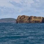 El Ilhéu Deitado, largo bloque de toba ocre con acantilados llenos de alvéolos, sobre un mar azul oscuro agitado, con la colina verde del Monte da Guia detrás y casas de Faial a lo lejos a la derecha