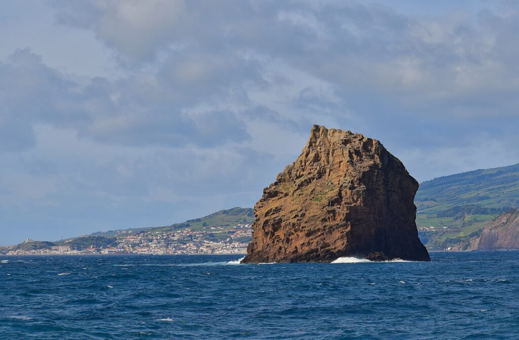 El Ilhéu em Pé, alta roca de toba ocre con estratos erosionados, se alza en un mar azul oscuro donde rompen algunas olas, frente a la ciudad blanca de Horta y las colinas verdes de Faial bajo un cielo nublado