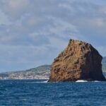 El Ilhéu em Pé, alta roca de toba ocre con estratos erosionados, se alza en un mar azul oscuro donde rompen algunas olas, frente a la ciudad blanca de Horta y las colinas verdes de Faial bajo un cielo nublado