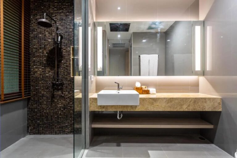 Una ducha junto a una pared de mosaico oscuro, al lado de un lavabo bajo un espejo iluminado