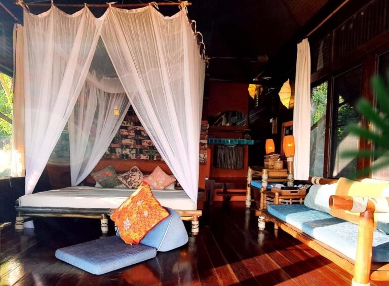 Una cabaña con una cama con mosquitero, un banco de bambú con cojines azules y un parquet oscuro