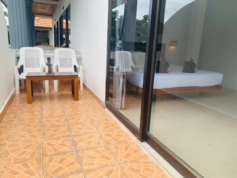Un balcón embaldosado con dos sillas blancas y una mesa baja, delante del ventanal de una habitación