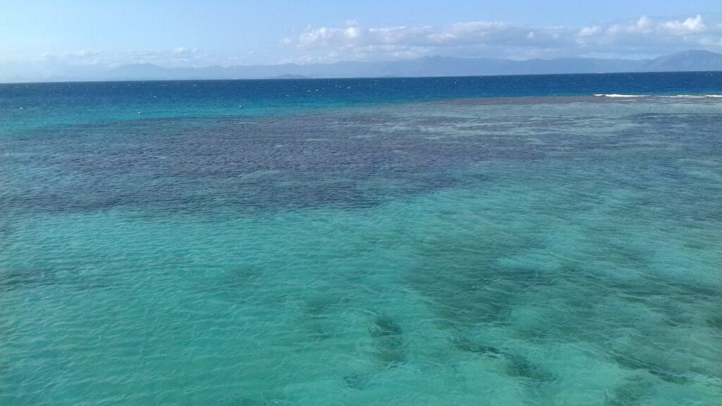 La laguna vista desde arriba, agua turquesa salpicada de macizos de coral oscuros, la barrera donde rompen las olas a la derecha y las montañas de la Grande Terre en el horizonte