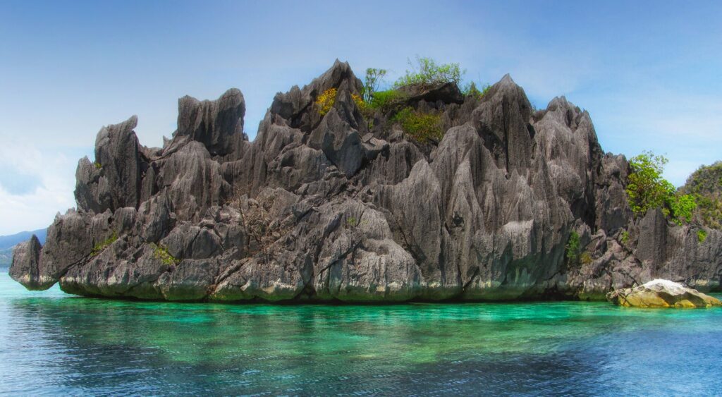 Un islote de caliza gris erizado de puntas, coronado por algo de vegetación, se alza sobre un agua turquesa transparente donde se adivina el coral, bajo un cielo azul.