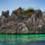 Un islote de caliza gris erizado de puntas, coronado por algo de vegetación, se alza sobre un agua turquesa transparente donde se adivina el coral, bajo un cielo azul.