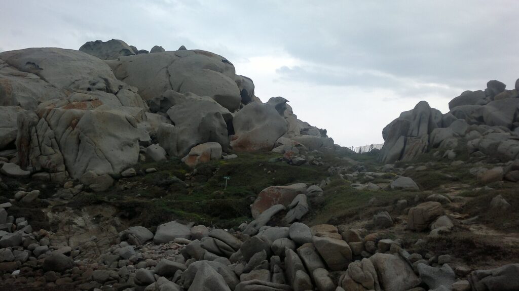 Montón de grandes bloques de granito gris redondeados y excavados por la erosión, manchados de líquenes naranjas, alrededor de un césped ralo donde un sendero bordeado por una valla de madera va hacia el mar, bajo un cielo cubierto.