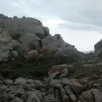 Montón de grandes bloques de granito gris redondeados y excavados por la erosión, manchados de líquenes naranjas, alrededor de un césped ralo donde un sendero bordeado por una valla de madera va hacia el mar, bajo un cielo cubierto.