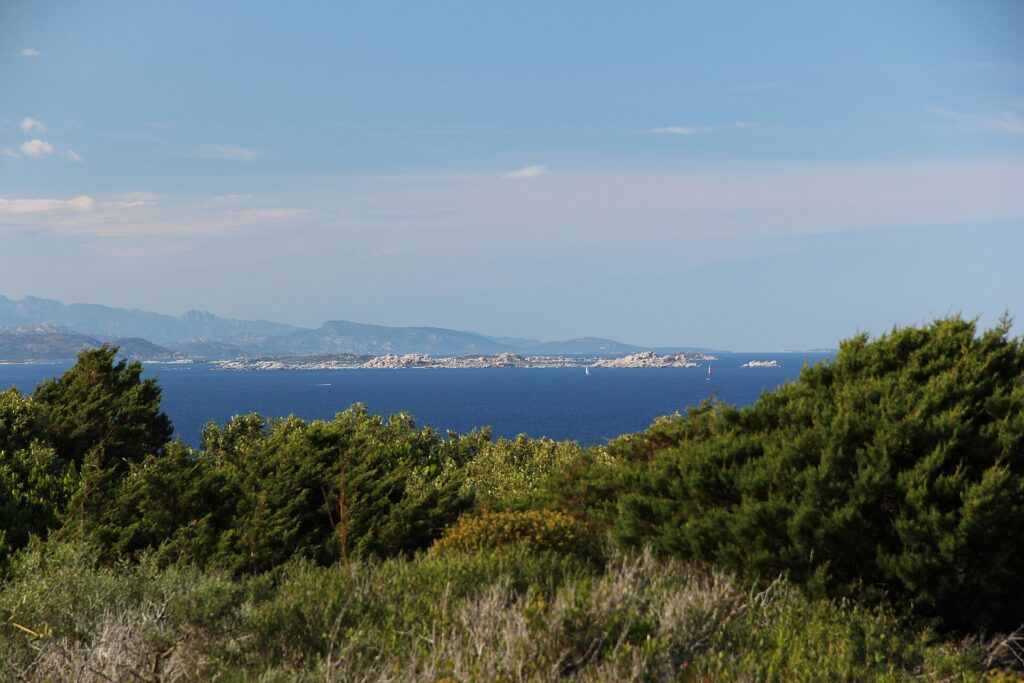 Vista desde el matorral de la costa de una isla larga y baja hecha de bloques de granito claro, sobre un mar azul oscuro, con un velero a sus pies y montañas azuladas en el horizonte.