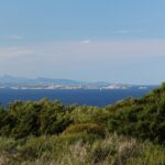 Vista desde el matorral de la costa de una isla larga y baja hecha de bloques de granito claro, sobre un mar azul oscuro, con un velero a sus pies y montañas azuladas en el horizonte.