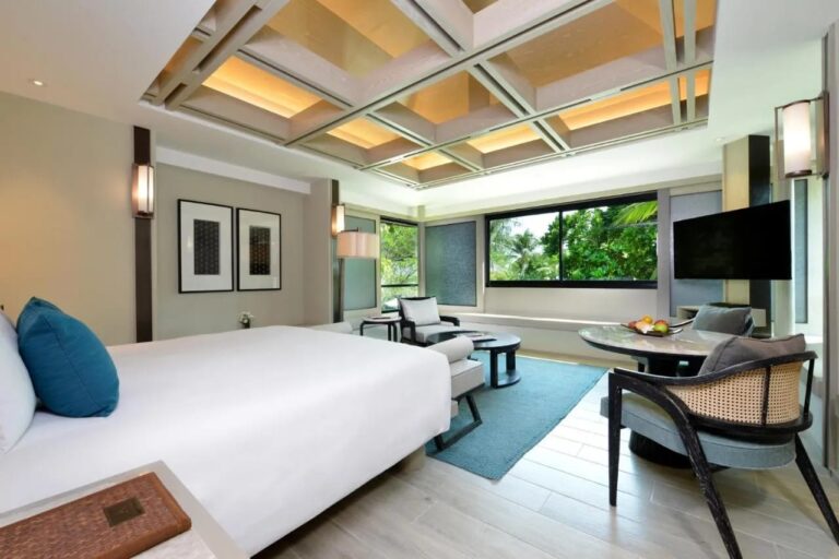 Una habitación con cama grande, techo artesonado, zona de estar y una ventana a las palmeras
