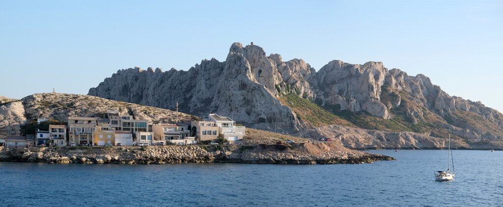 Las casas del pueblo de Les Goudes alineadas a ras de una costa rocosa, frente a una alta cresta de caliza blanca de picos dentados, bajo un cielo azul, con un velero fondeado en un mar en calma.