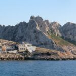 Las casas del pueblo de Les Goudes alineadas a ras de una costa rocosa, frente a una alta cresta de caliza blanca de picos dentados, bajo un cielo azul, con un velero fondeado en un mar en calma.