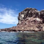 Las rocas rojizas de Los Islotes bajo un cielo azul, con lobos marinos tumbados a ras del agua y un acantilado agrietado encima
