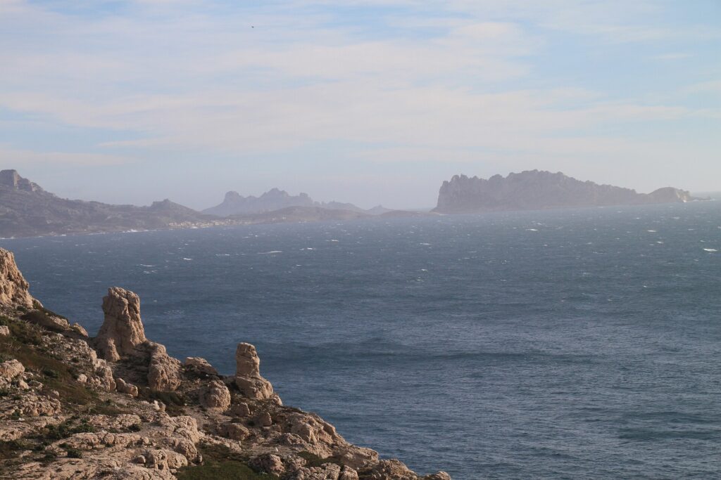 Desde las rocas calizas del Frioul en primer plano, vista de un mar agitado y cubierto de espuma hasta la isla Maïre y la costa de Les Goudes, recortadas en la bruma.
