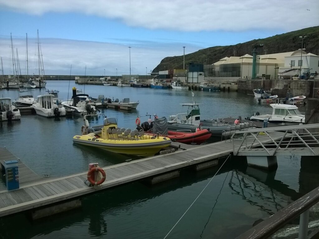 Lanchas semirrígidas, una amarilla con filas de asientos y una roja, amarradas al pantalán de la marina de Vila do Porto, con veleros y barcos de pesca al fondo bajo un acantilado verde y un cielo nublado