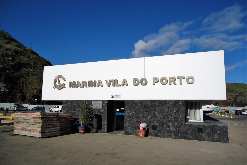 El edificio de recepción de la marina de Vila do Porto, un gran rótulo blanco con letras metálicas sobre un muro de basalto negro, bajo un cielo azul, con un acantilado verde a la izquierda