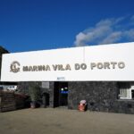 El edificio de recepción de la marina de Vila do Porto, un gran rótulo blanco con letras metálicas sobre un muro de basalto negro, bajo un cielo azul, con un acantilado verde a la izquierda