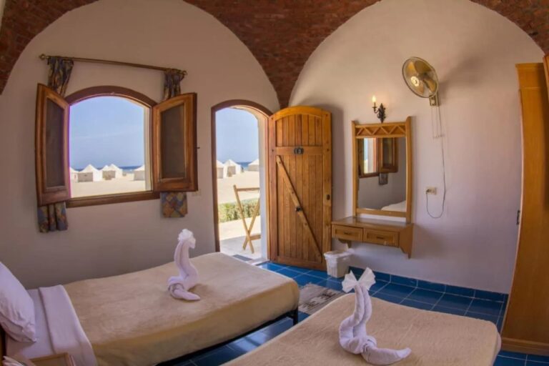 Una habitación con dos camas abierta a la playa, con carpas blancas y el mar al fondo