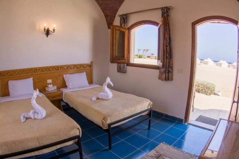 Una habitación con dos camas bajo una bóveda de ladrillo, con la puerta abierta a la arena