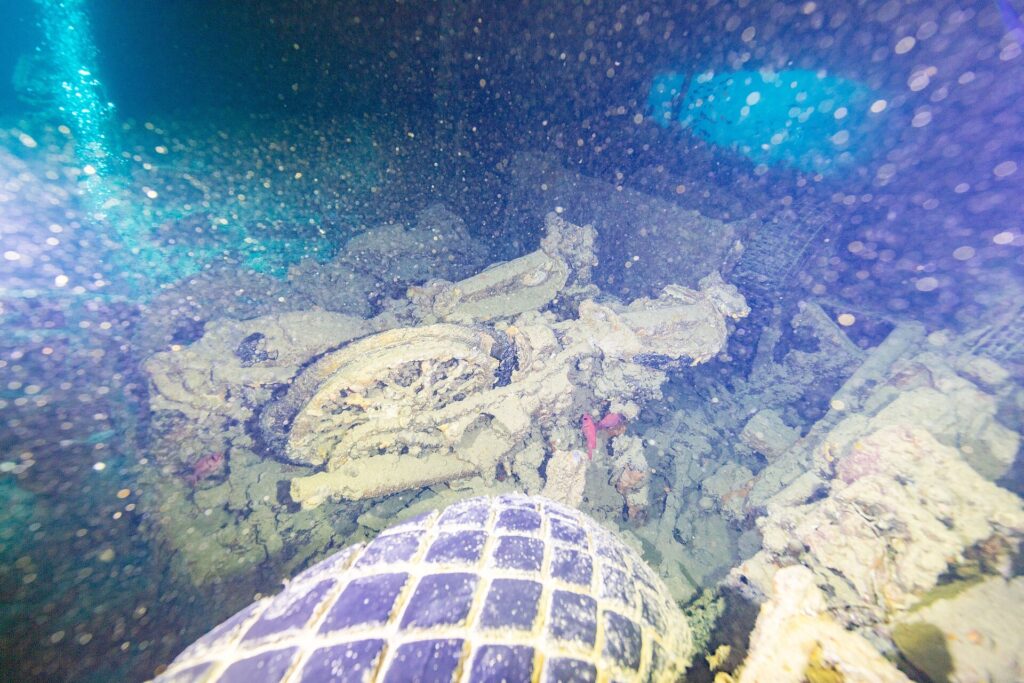 En una bodega oscura del Thistlegorm, una moto volcada detrás de un neumático de camión, iluminada con linterna