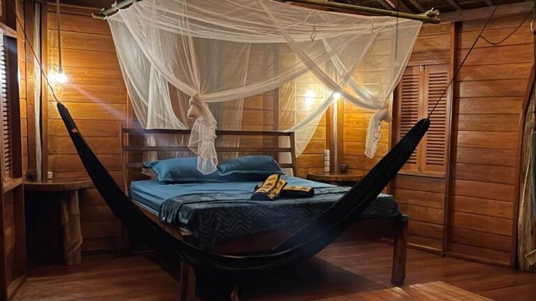 Una habitación con paredes de madera iluminada por lámparas, con una cama con mosquitero y una hamaca negra